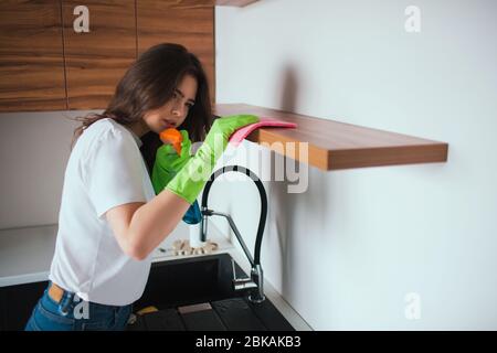 Young woman in kitchen during quarantine. Girl cleaning shelf surface carefully. Using cloth and cleaning spray. Wear green gloves around hands. Stock Photo
