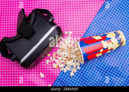 Virtual reality glasses and overturned bucket of popcorn on pink blue table. Stock Photo