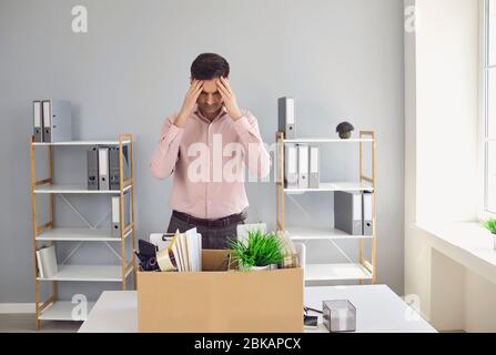 Unemployment. Dismissal. Dismissed businessman is upset with a cardboard box holding his head in his hands while sitting in a company office. Stock Photo