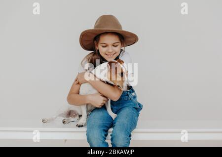 Little adorable girl wars fashionable hat and denim overalls, embraces favorite dog with love, have fun and play together, sit on desk against white b Stock Photo