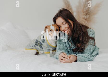 Photo of beautiful brunette woman in knitted sweater lies together wih dog on soft bed, enjoys spending time with favorite pet, cares about animals, s Stock Photo