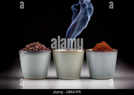 3 rustic coffee cups with coffee grounds, smoking coffee, and ground coffee. Standing on a white reflective table fading into a black background Stock Photo