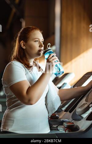 pensive overweight girl drinking from sports bottle while standing at treadmill