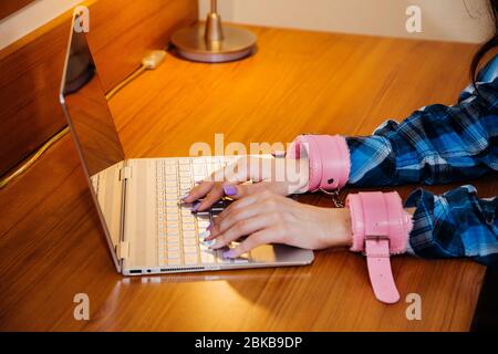 Hands in pink handcuffs are typing on the keyboard of a thin laptop; symbolic for being stuck to the computer during quarantine or stress or addiction Stock Photo