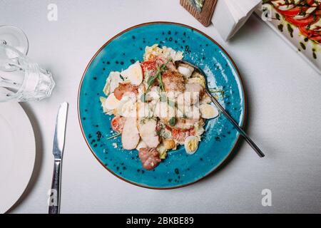 salad with different meat, tomatoes and eggs, all sprinkled with parmesan on a blue plate which stands on a white tablecloth Stock Photo