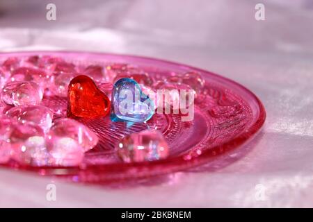 A red and a blue glass heart surrounded by pink hearts on a pink glass plate with ornament pattern. Wedding card background. Stock Photo