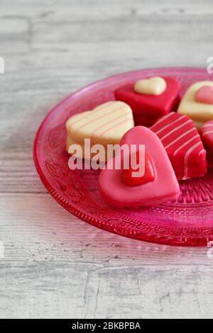Heart shaped colorful chocolates (petit fours) on a pink glass plate with ornament pattern. Light wooden table background. Stock Photo