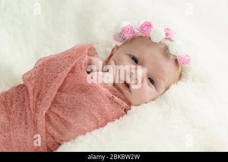 A small newborn girl in a pink diaper and a wreath of rose flowers lies on a white blanket and looks at the camera. Close-up portrait Stock Photo