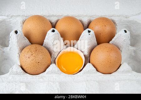 Fresh raw chicken eggs in a paper carton. One egg is half broken and contains a yolk. Close-up shot, top-side view. Stock Photo