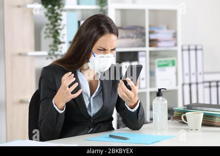 Angry executive woman looking smart phone avoiding covid-19 with mask sitting on a desk at office Stock Photo