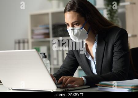 Concentrated executive woman working late on laptop avoiding covid-19 with mask sitting at night in the office Stock Photo