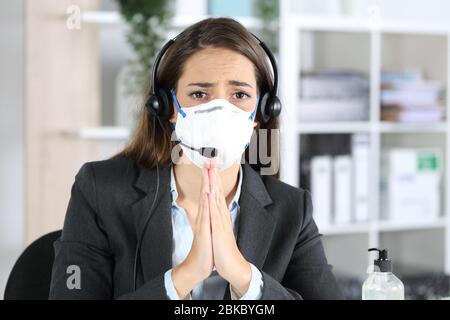Front view of sad telemarketer woman avoiding covid-19 with mask begging looking camera sitting at the office Stock Photo