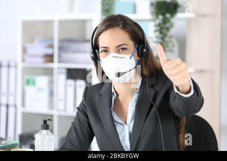 Happy telemarketer woman with thumbs up looking camera avoiding covid-19 with mask sitting in the office Stock Photo