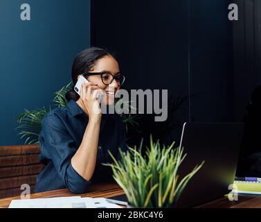Smiling woman sitting at desk looking at laptop screen and talking over mobile phone Stock Photo