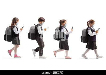 Full length profile shot of schoolchildren with protective face masks walking in line and holding mobile phones isolated on white background Stock Photo