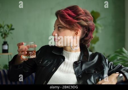 Young sad woman with dark pink hair with glam rock style look is drinking whisky from a glass with abomination grimace on the face, female alcoholism Stock Photo