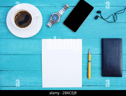 Mockup of a white cup of coffee, black notebook, phone, black headphone, man watch, yellow pen, and white empty sheet on turquoise wood desk. Stock Photo