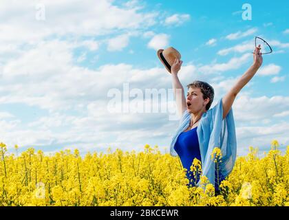 Happy woman wearing blue top and jeans  raised her hands high against blu sky and yellow rapeseed fields Stock Photo