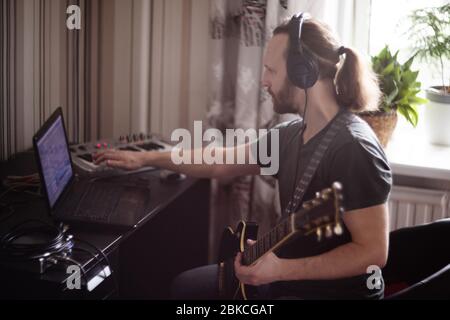 Bearded man musician playing music and composing a song with electric guitar piano and laptop computer while sitting in living room, corona virus quar Stock Photo