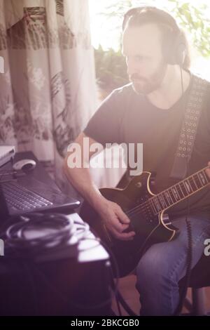 Bearded man musician playing music and composing a song with electric guitar piano and laptop computer while sitting in living room, corona virus quar Stock Photo
