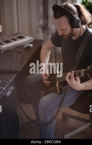 Bearded man musician playing music and composing a song with electric guitar piano and laptop computer while sitting in living room, corona virus quar Stock Photo