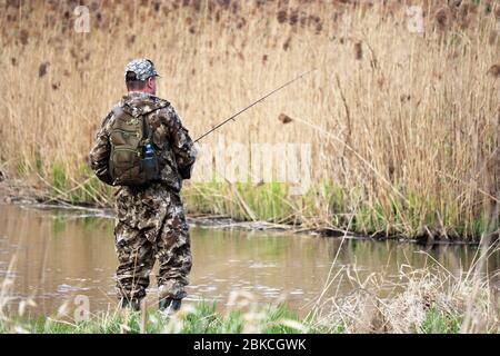 Fisherman in hunting clothes standing with a fishing rod. Man angling in spring on the river coast overgrown with reeds Stock Photo