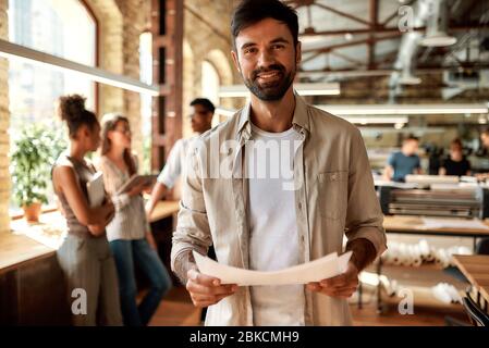 Great working day! Young bearded man in casual clothes holding documents and looking at camera with smile while standing in the modern office Stock Photo