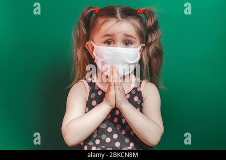 Caucasian sick little girl in medical mask during coronavirus epidemic prays on green background closeup 2021 Stock Photo
