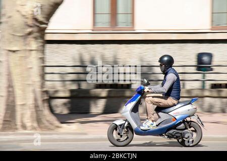 Belgrade, Serbia - April 23, 2020: A young man wearing helmet, face mask and sunglasses riding a scooter motorbike on city street Stock Photo