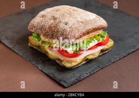 A sandwich on a tray. Sandwich with meat and vegetables on a black granite stand. Special square roll for sandwiches. Stock Photo