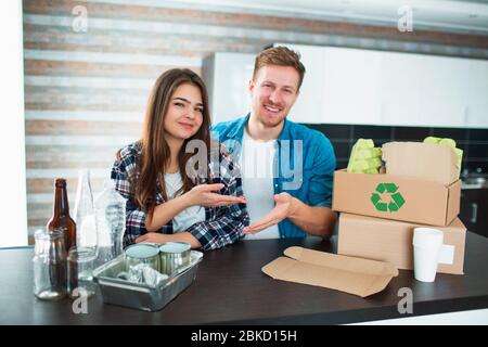 A young couple sorts trash in the kitchen. Young man and woman are sorting recyclables in the kitchen. There is cardboard, paper, iron, plastic and Stock Photo