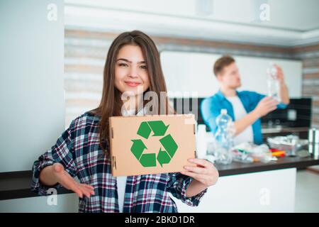 Young family sorts materials in the kitchen for recycling. Recyclable materials must be separated. Wife is holding a cardboard box with a green Stock Photo