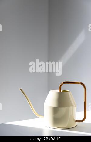 Close up of elegant white metallic watering can with long thin spout and yellow thin circular handle on the table, wall on background, indoors. Sunlig Stock Photo