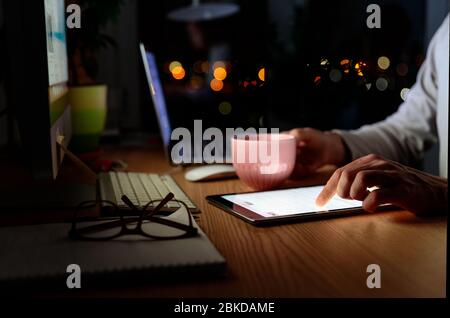 Freelancer man holds a mug of tea, scrolling through social networks, news search on digital tablet late at night during a break in work. Selective fo Stock Photo