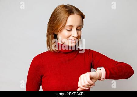 Happy stylish business woman in red turtleneck looking at smartwatch on her wrist, checking message on screen, checking the time. Isolated on grey bac Stock Photo
