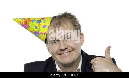 Portrait of drank sleepy young clerk man with a hangover giving thumbs up, smiles. Emotions. Disheveled guy businessman in suit and festive cap looks at camera. White background. Office birthday party Stock Photo