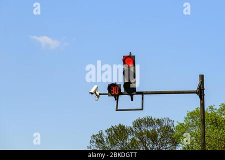 Red traffic light with digital timing countdown and traffic security camera surveillance (CCTV) installed on a pole above a roadway on a sunny day. Stock Photo