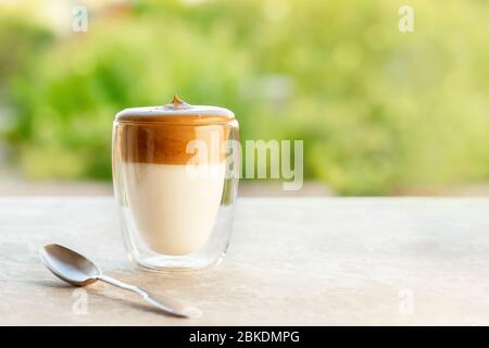 Dalgona frothy coffee in glass with spoon on table on green background. Trend korean Iced  latte coffee drink with foam of instant coffee with copy sp Stock Photo