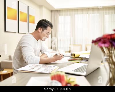 young asian man design professional sitting at kitchen counter working from home (artwork in background digitally altered) Stock Photo