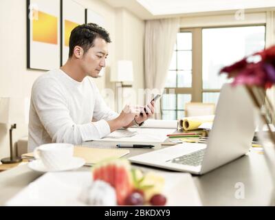 young asian man design professional working from home sitting at kitchen using cellphone (artwork in background digitally altered) Stock Photo