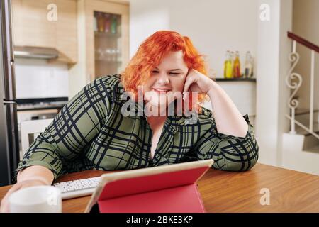 Happy young plus size woman sitting at kitchen table and watching tv series on tablet computer Stock Photo