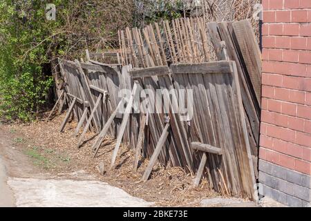 Old rickety wooden fence with props. Brick wall nearby. Green bushes on the background. Horizontal. Stock Photo