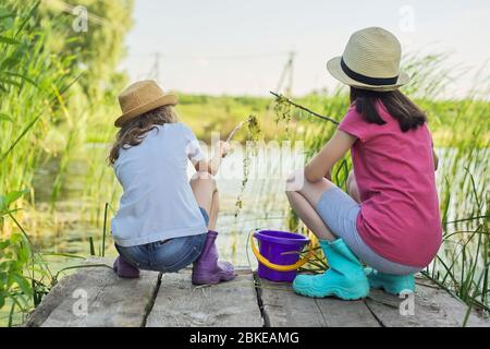 Kids girls sitting on wooden pier, catching water snails in bucket Stock Photo