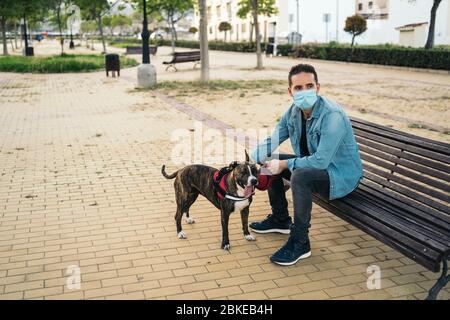 Young man in medical mask sitting on a bench in the park with his dog. Covid-19 coronavirus pandemic. Active quarantine life in the Corona outbreak. Stock Photo