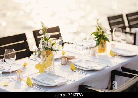 Wedding dinner table reception. Rectangular tables with white tablecloth, floral arrangements lemons in vases. Yellow paper butterflies are scattered Stock Photo