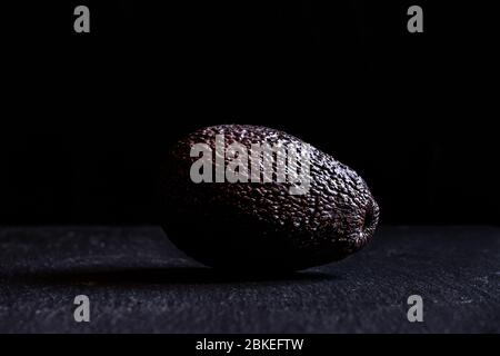 avocado on dark black background chiaroscuro closeup with copy space whole oval dark green ripe Stock Photo