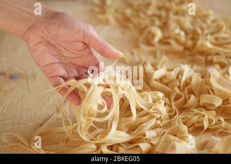 Preparing fresh homemade pasta: close-up view of woman's hand pulling up some fresh tagliatelle on wooden work table in natural sunlight Stock Photo