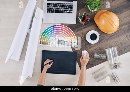 Interior designer working on graphic tablet at desk with color palette, blueprints and cup of coffee, view from above Stock Photo