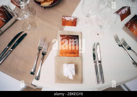 Close-up of a wedding dinner table. Brown napkins with an enclosed menu, forks and knives, wine glasses against the backdrop of a cream tablecloth Stock Photo