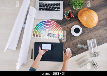 Interior designer checking house blueprint on screen of smartphone when making project on computer, view from above Stock Photo